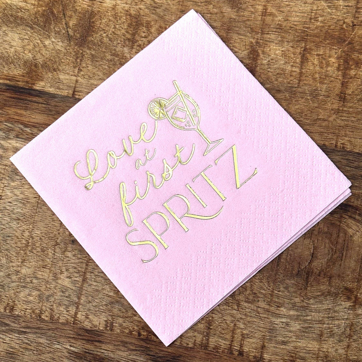 A stack of light pink cocktail napkins with gold foil stamping that reads Love at first SPRITZ alongside a cocktail illustration, set on a wood background.