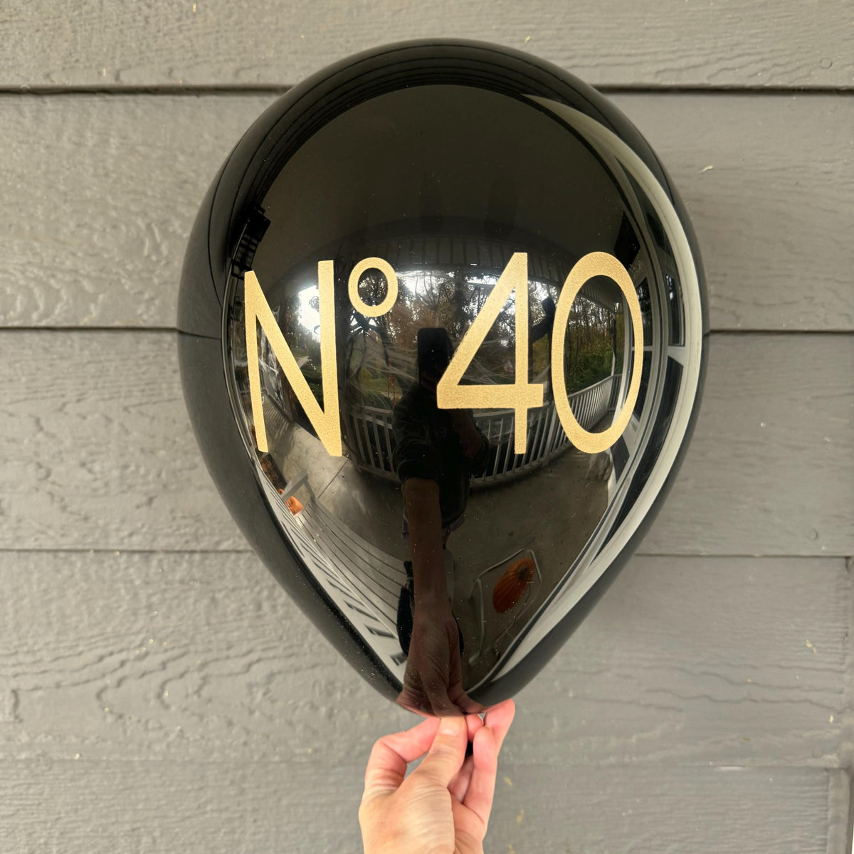Close-up of a hand holding a black No 40 birthday balloon with gold lettering against gray siding.