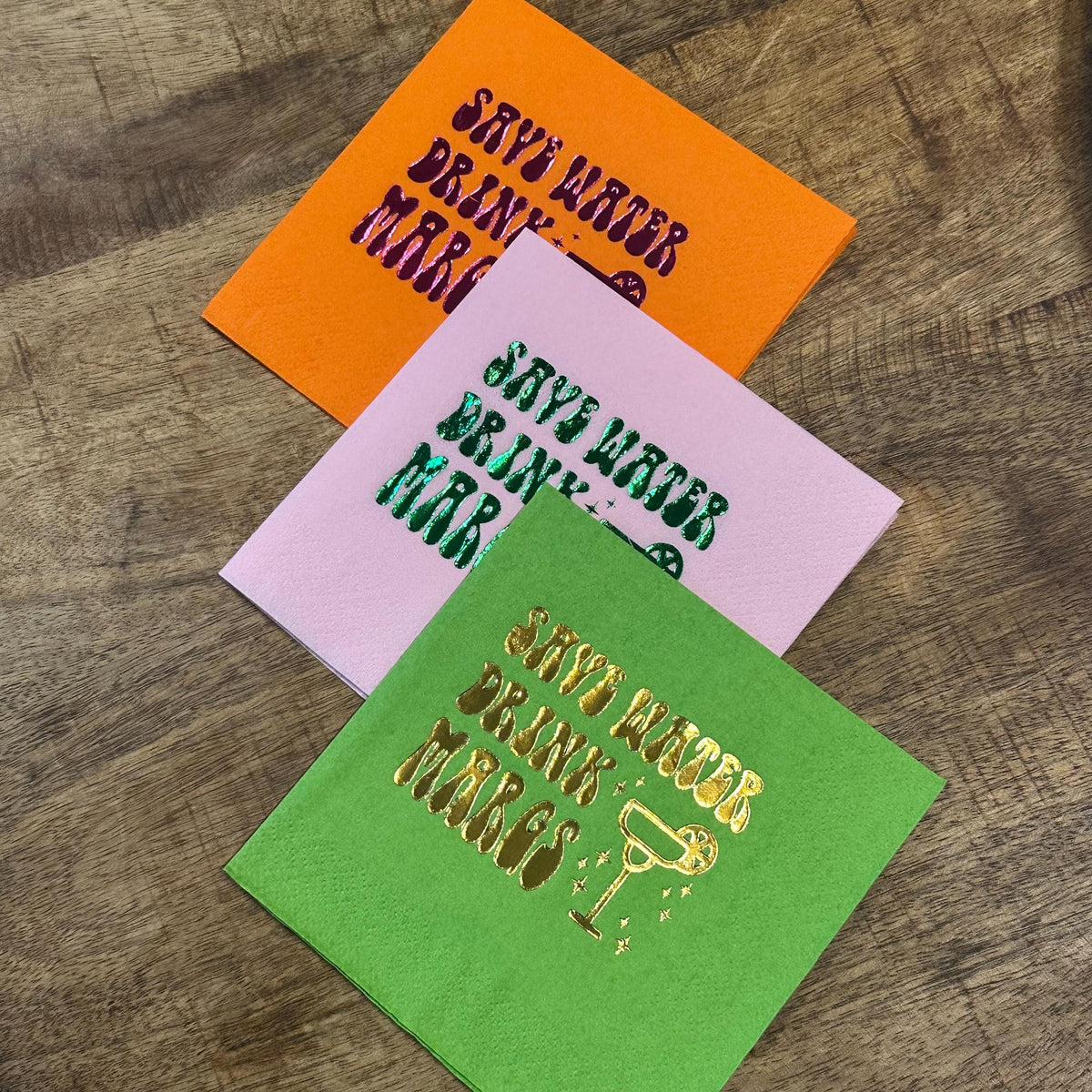 Three folded party napkins stacked diagonally on a wooden table, featuring an orange napkin on top, a pink napkin in the middle, and a green napkin at the bottom with metallic foil text.