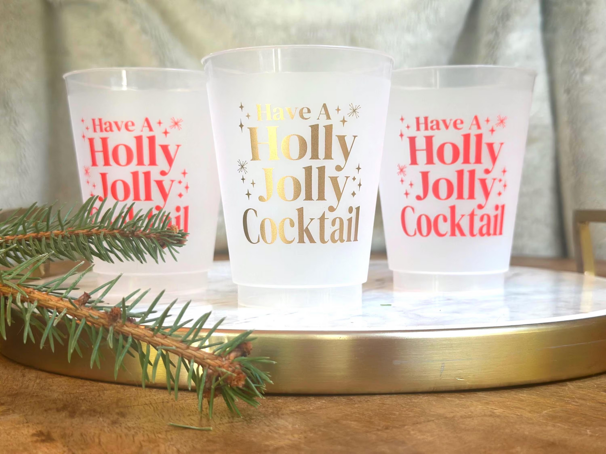 Frosted plastic cups with the festive phrase “Have a Holly Jolly Cocktail” printed in gold and red lettering. The cups are displayed on a round marble tray with a gold rim, accented by a pine branch in the foreground — a cheerful holiday drinkware setup perfect for Christmas parties.