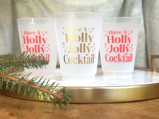 Frosted plastic cups with the festive phrase “Have a Holly Jolly Cocktail” printed in gold and red lettering. The cups are displayed on a round marble tray with a gold rim, accented by a pine branch in the foreground — a cheerful holiday drinkware setup perfect for Christmas parties.