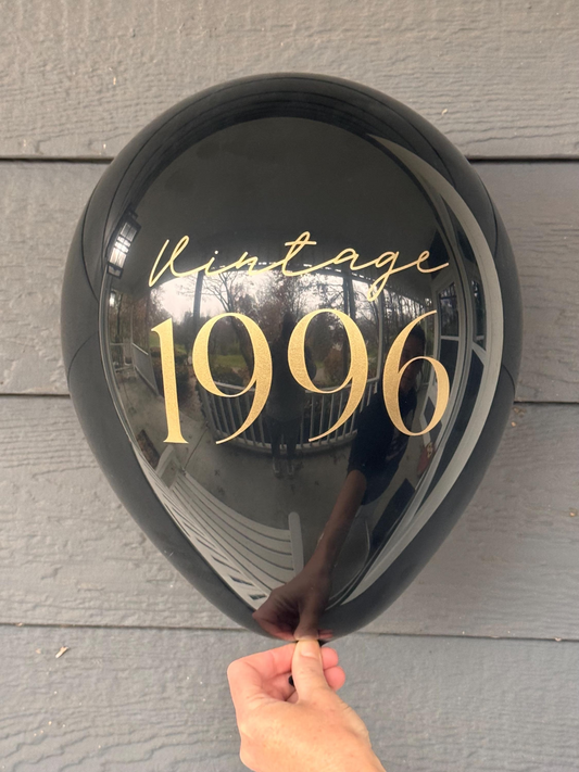 Black balloon with 'Vintage 1996' text held against a wooden surface