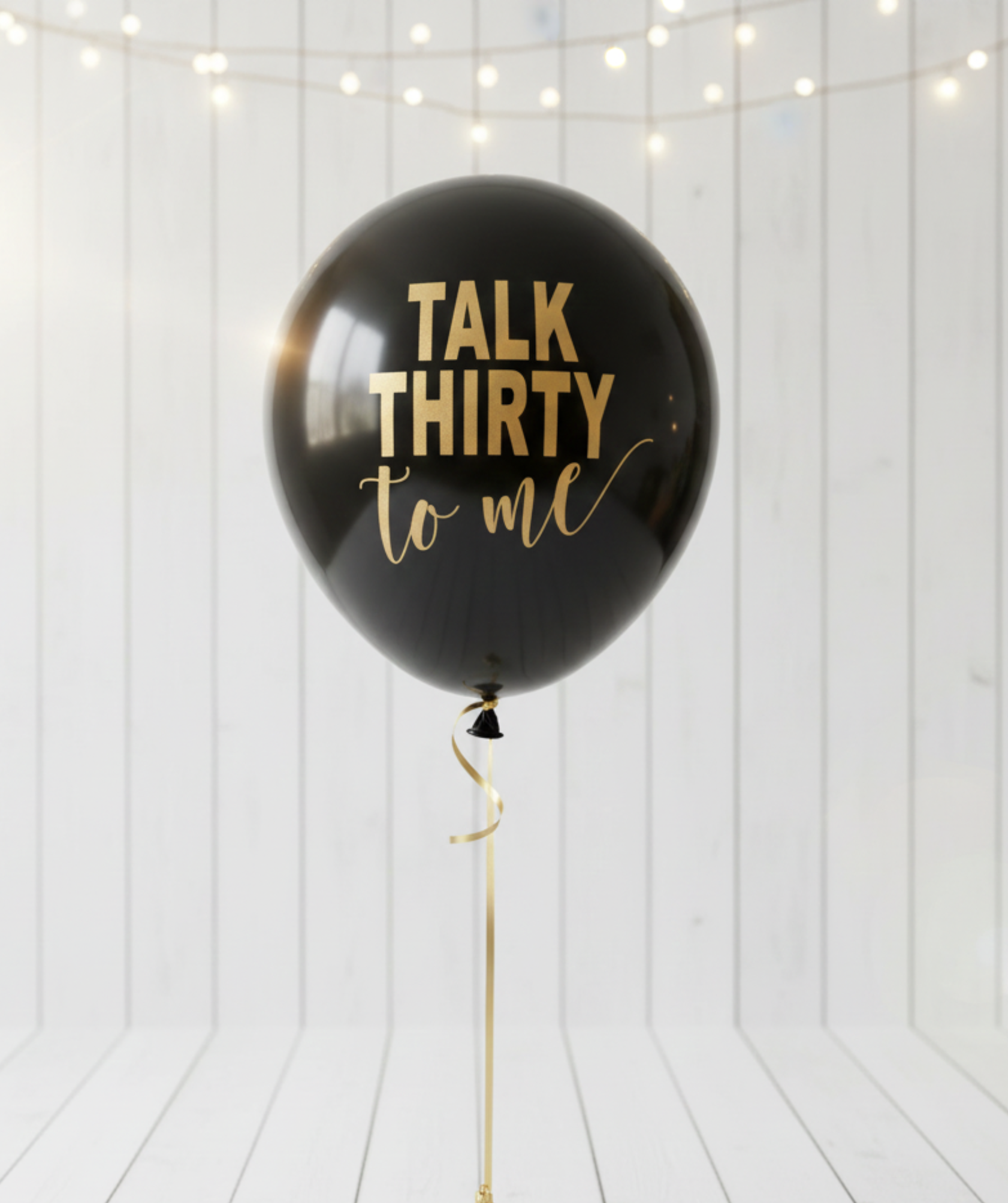 Black balloon with 'Talk Thirty to me' text on a white wooden floor with string lights.