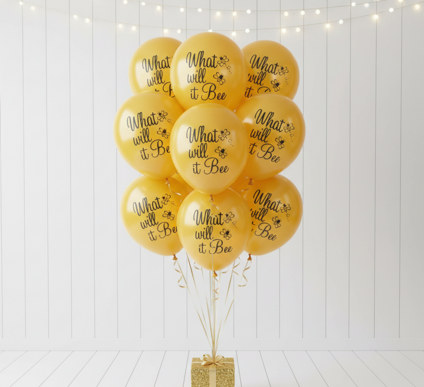 Yellow balloons with 'What will it be?' text and a gold gift box on a white wooden background.
