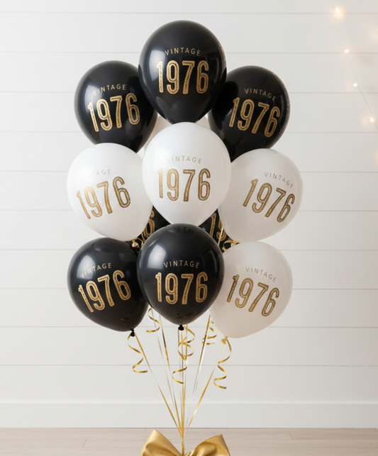 Decorative balloons with 'Vintage 1976' text on a stand with a gold bow and gift box against a white wooden panel background.