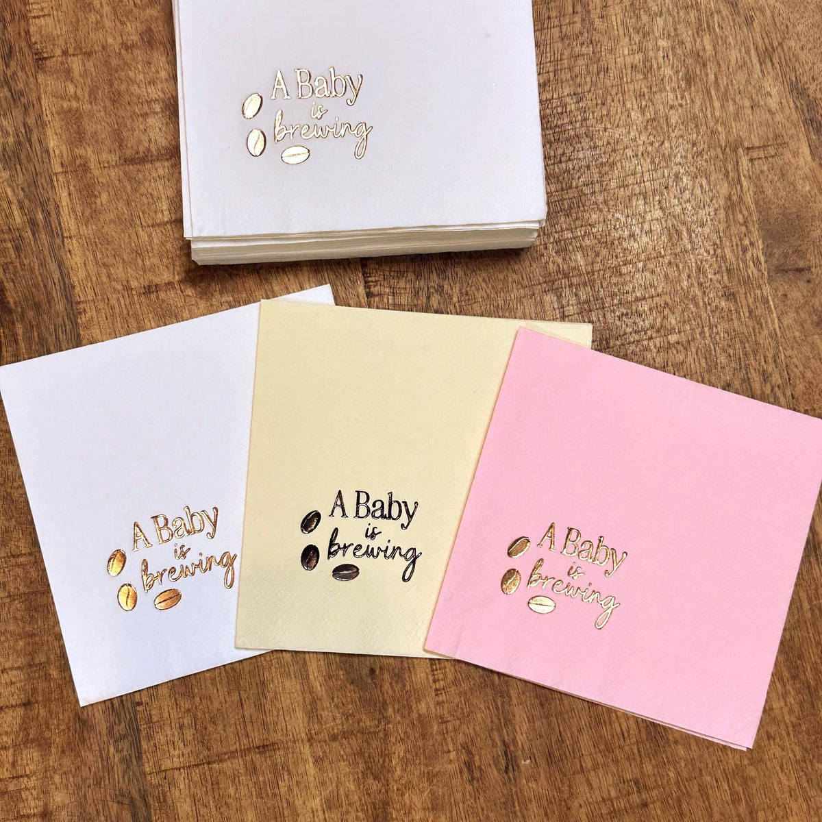 Three square napkins, one white, one cream, and one pink, each foil-stamped with "A Baby is Brewing" and three coffee beans in gold, with a stack of white napkins behind them.