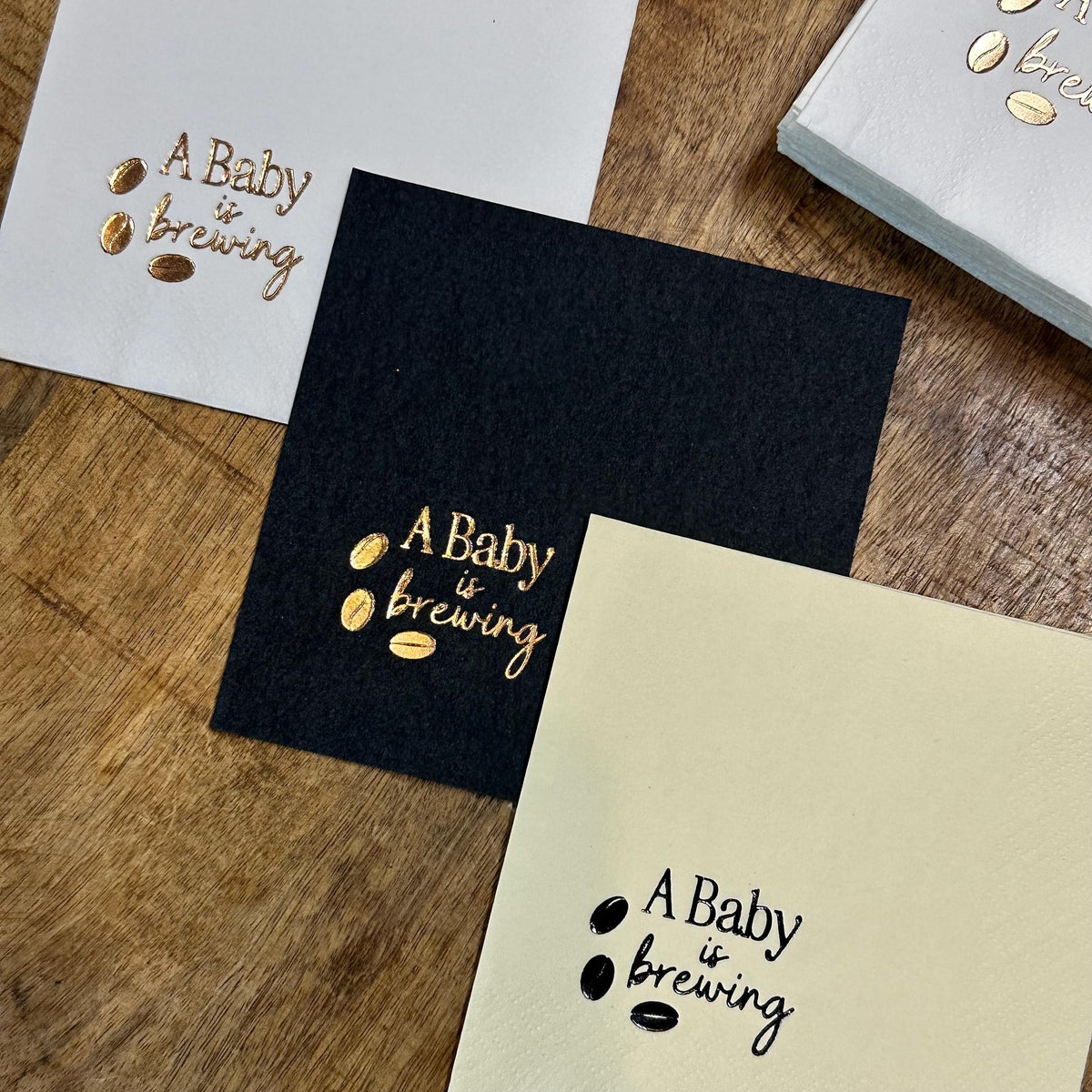 Three square paper napkins, one white, one black, and one cream, each foil-stamped with "A Baby is brewing" and three coffee beans in metallic gold.