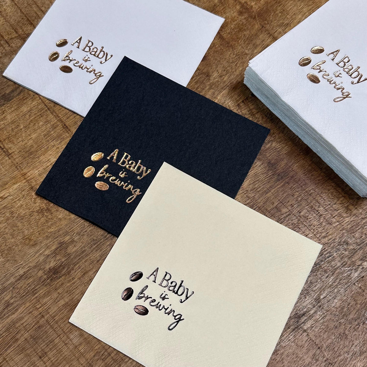 Three napkins, white, black, and cream, each foil-stamped with "A Baby is brewing" and coffee beans, with a stack of white napkins in the background.