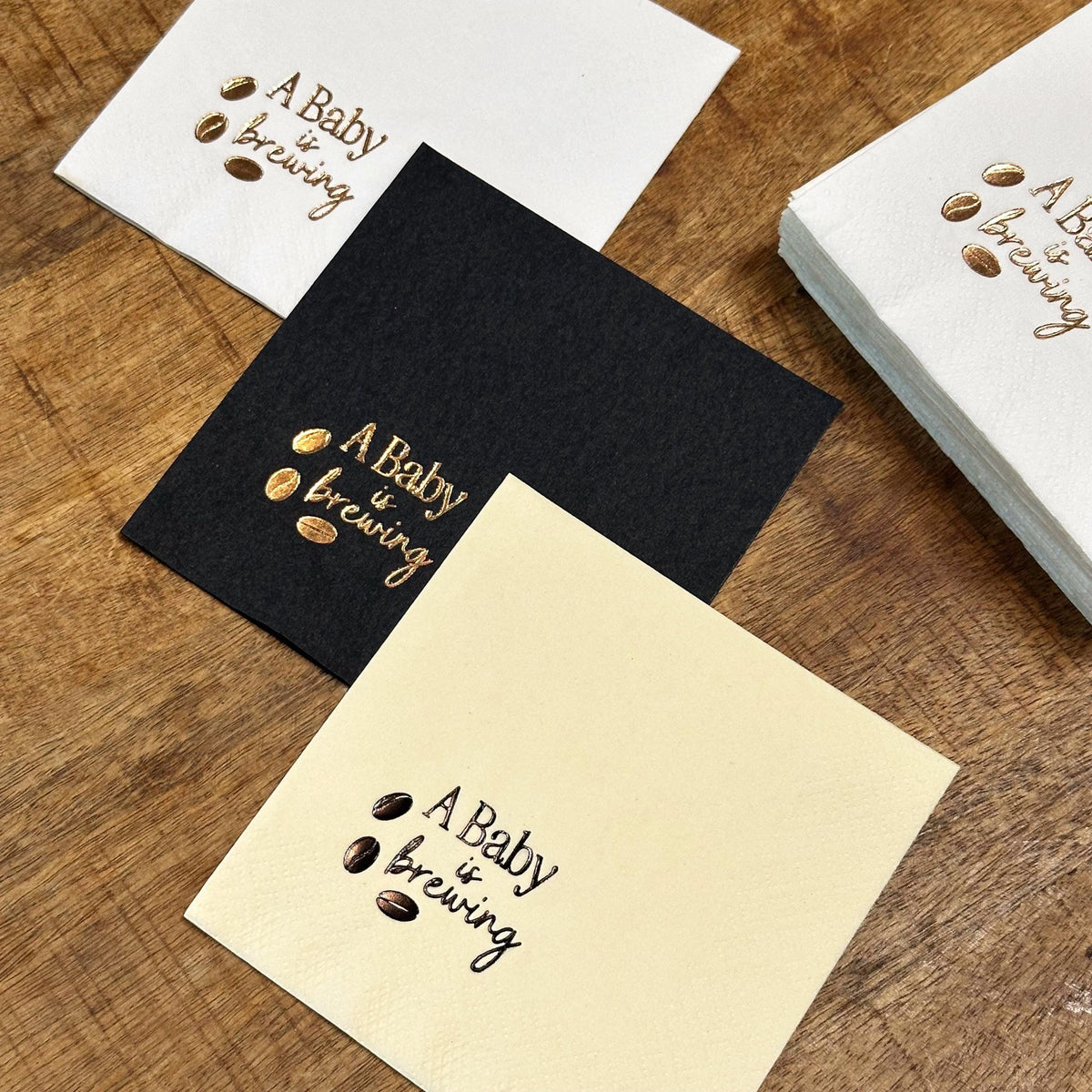 Three napkins, white, black, and cream, each foil-stamped with "A Baby is brewing" and coffee beans, with a stack of white napkins to the right.