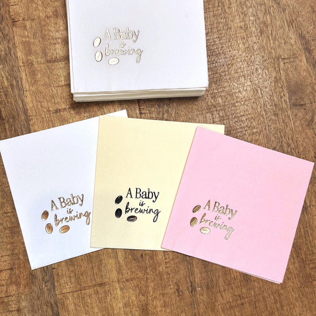Three square paper napkins, white, cream, and pink, each foil-stamped with "A Baby is brewing" and three coffee beans in metallic gold, with a stack of white napkins behind them.