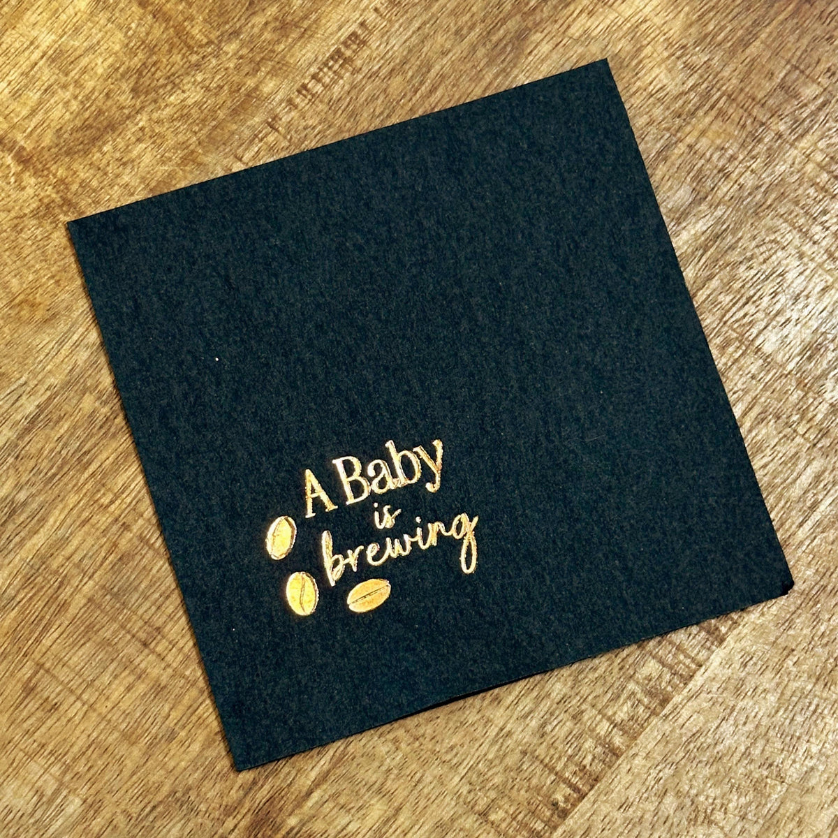 A single black square napkin with "A Baby is Brewing" and three coffee beans foil-stamped in gold.