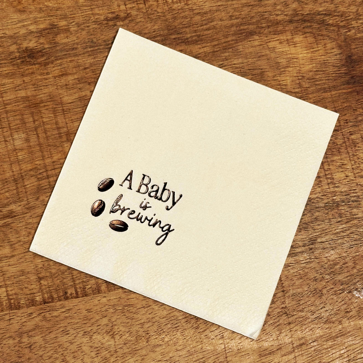 A single cream-colored square napkin with "A Baby is Brewing" and three coffee beans printed in dark brown.