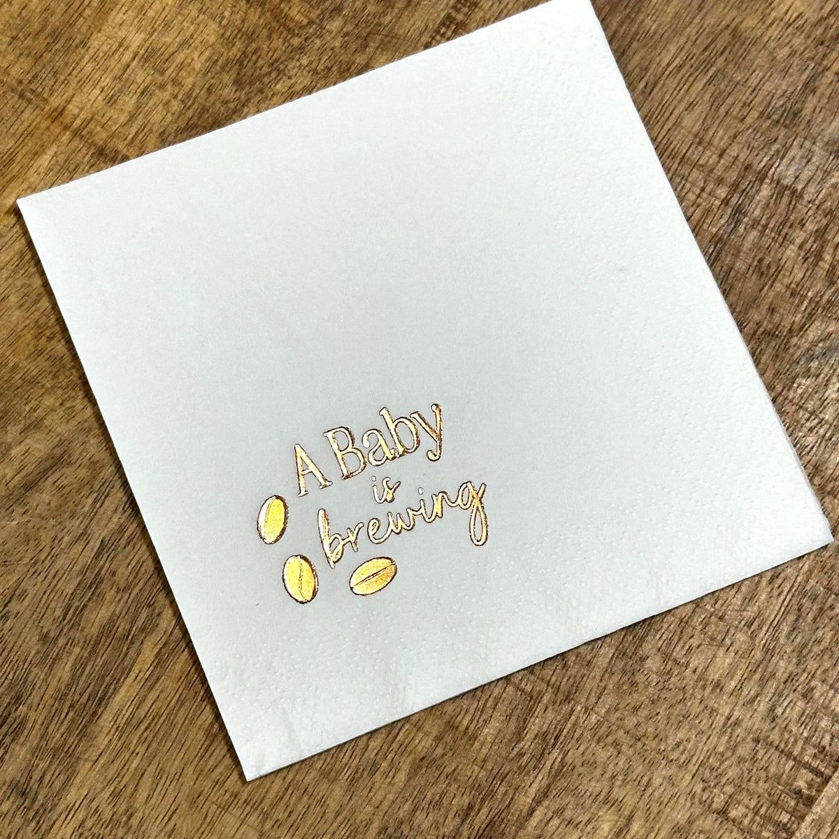A single white square napkin with "A Baby is Brewing" and three coffee beans foil-stamped in gold.