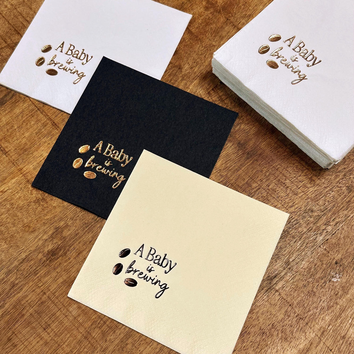 Three square napkins, one white, one black, and one cream, each printed with "A Baby is Brewing" and three coffee beans in dark brown, with a stack of white napkins behind them.