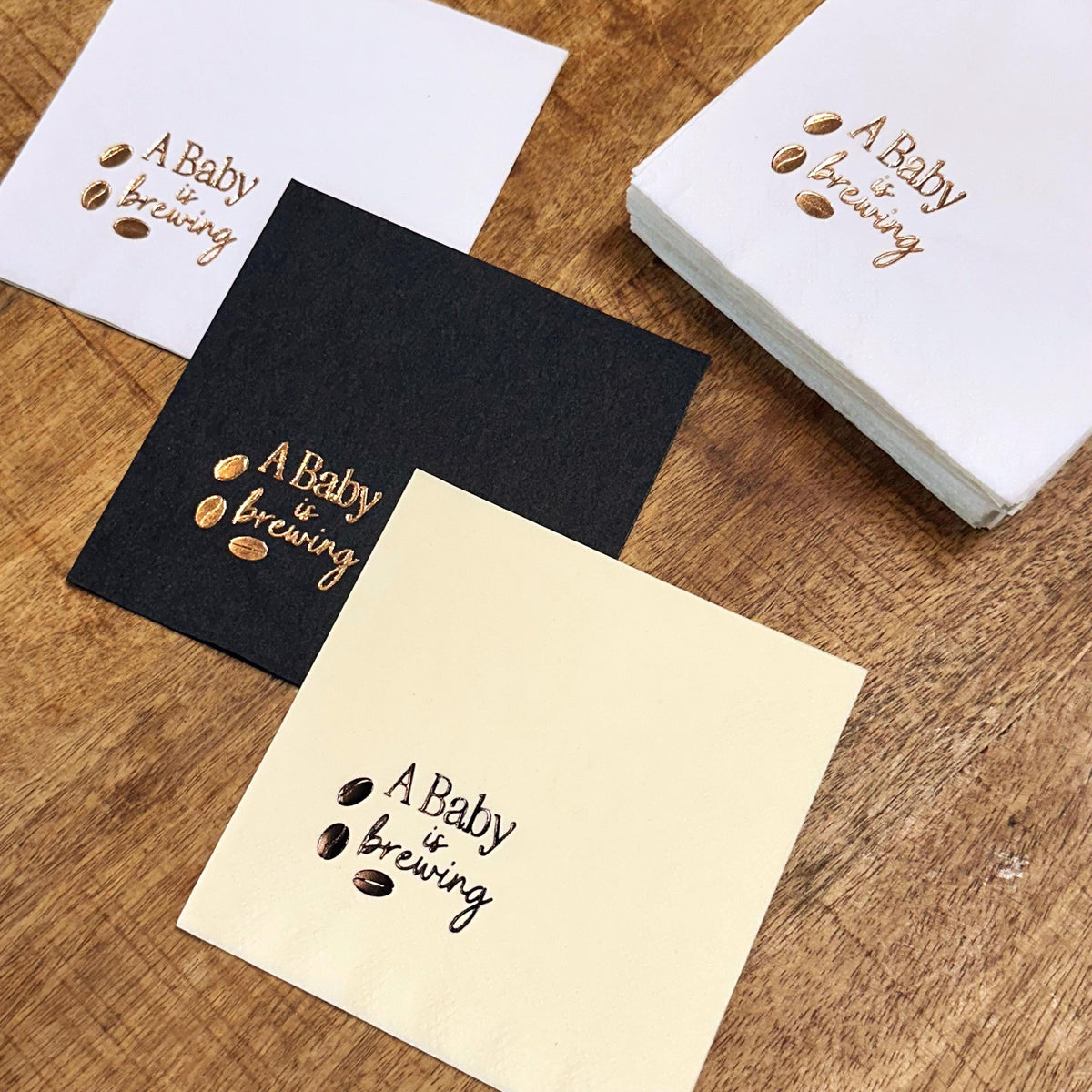 Three square napkins, one white, one black, and one cream, each printed with "A Baby is Brewing" and three coffee beans in copper, with a stack of white napkins behind them.