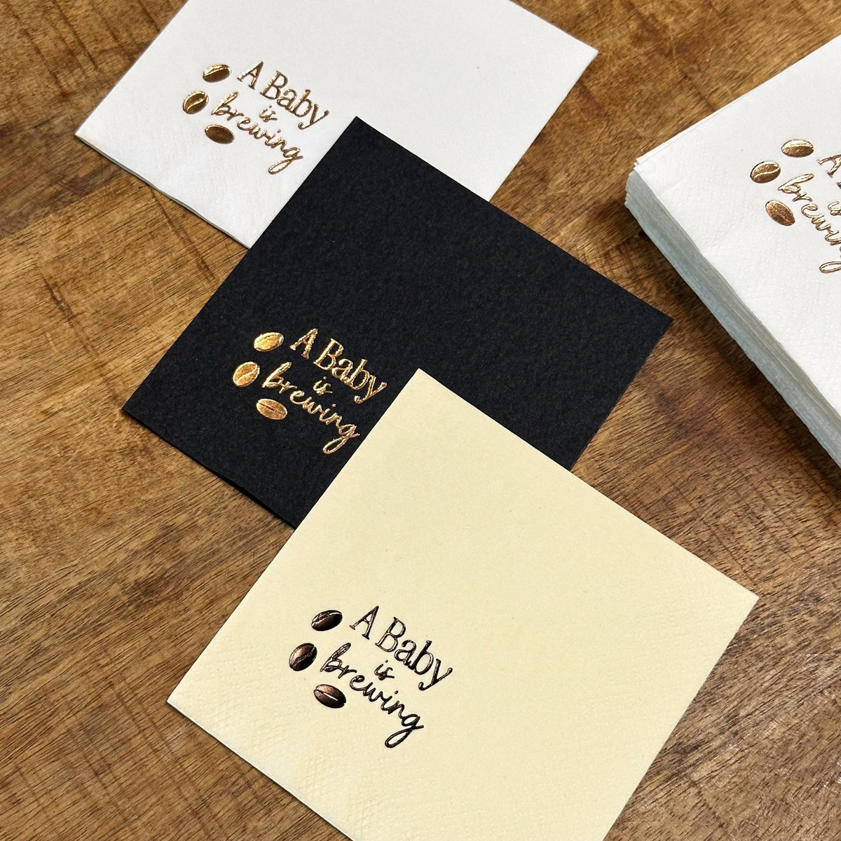 Three square napkins, one white, one black, and one cream, each foil-stamped with "A Baby is Brewing" and three coffee beans in gold, with a stack of white napkins behind them.