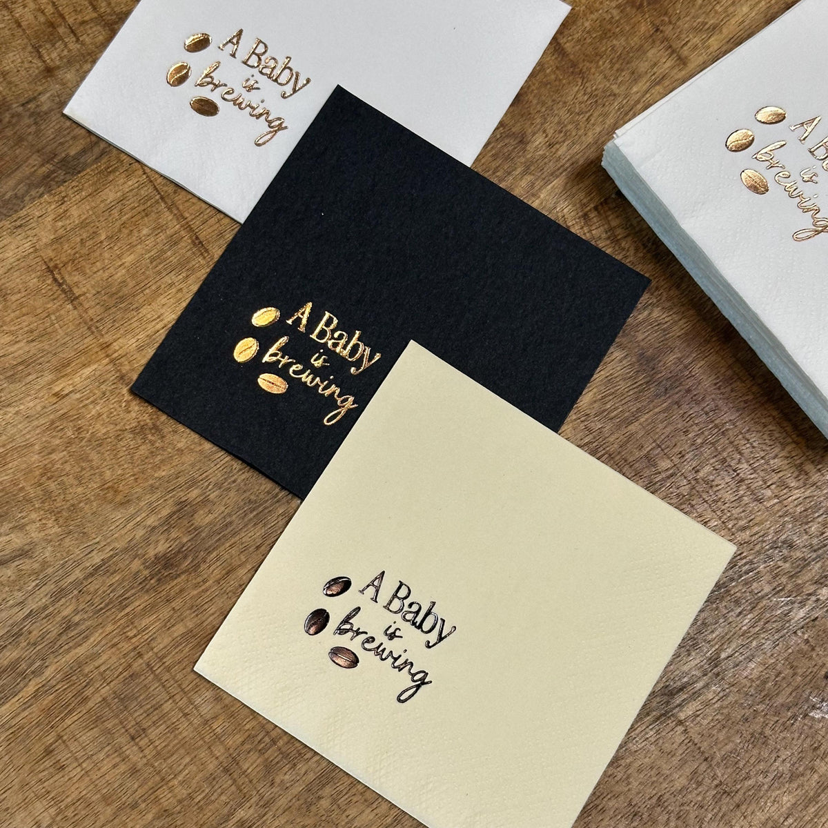 Three square napkins, one white, one black, and one cream, each foil-stamped with "A Baby is Brewing" and three coffee beans in gold, with a stack of white napkins behind them.