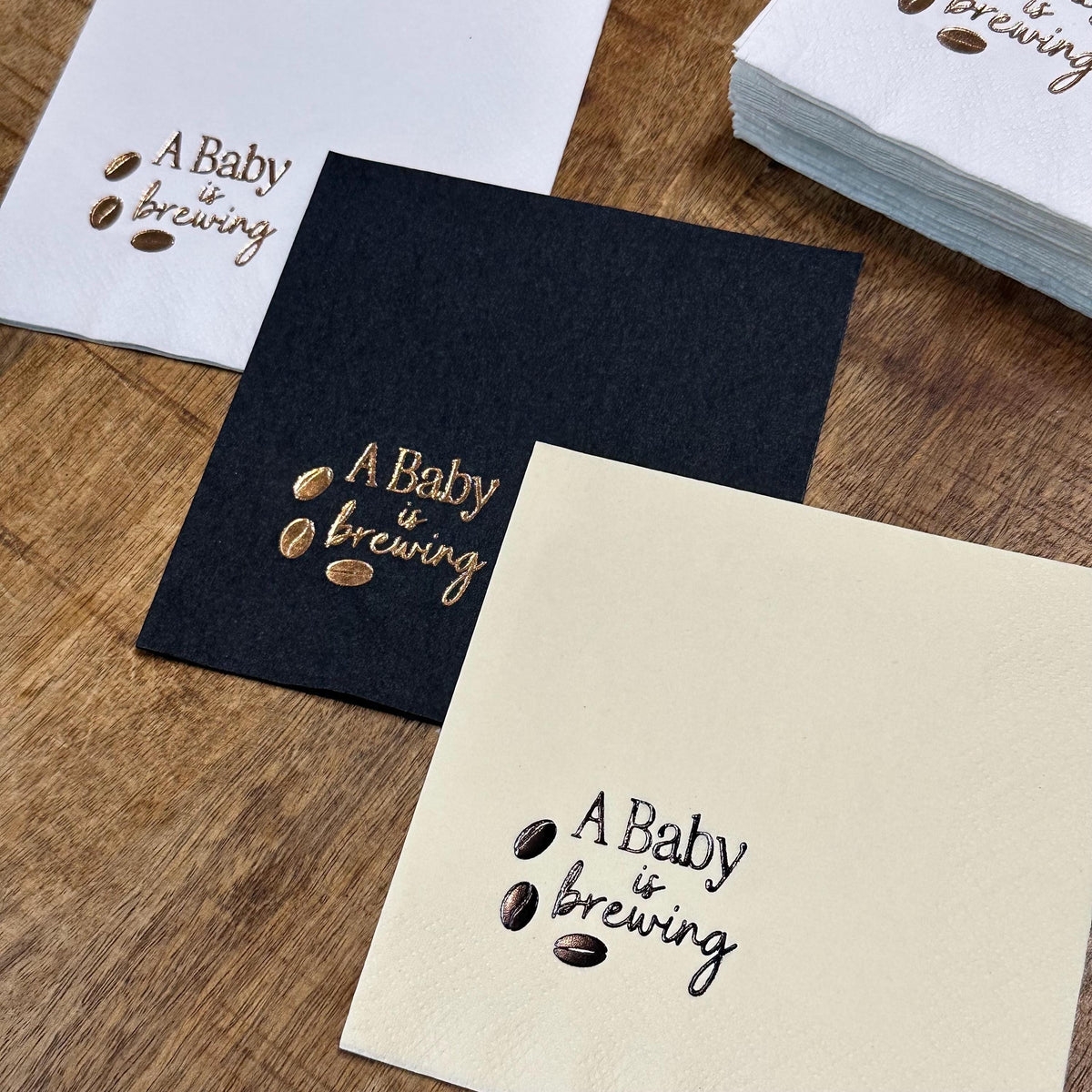 Three square napkins, one white, one black, and one cream, each printed with "A Baby is Brewing" and three coffee beans in dark brown, with a stack of white napkins behind them.