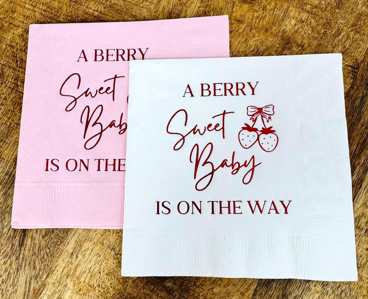 Two square cocktail napkins on a wooden surface, one pink and one white, both printed with the text A Berry Sweet Baby Is On The Way.