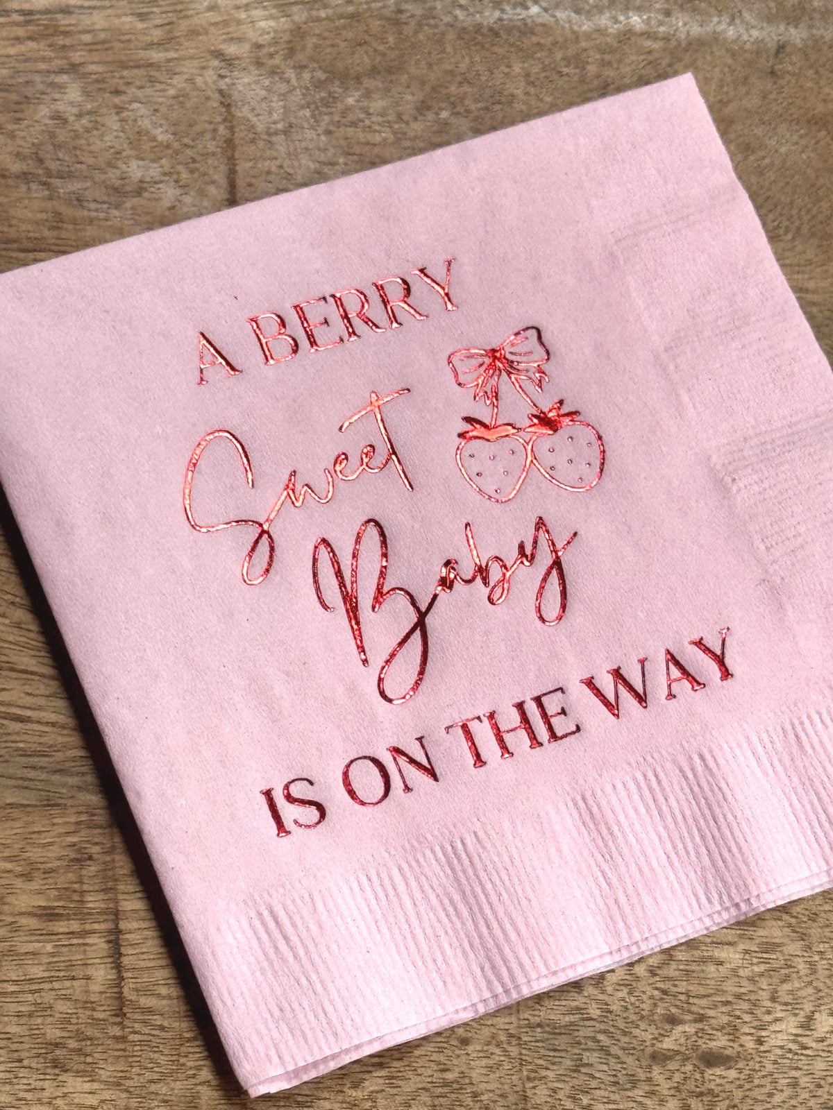 A light pink cocktail napkin on a wooden table featuring metallic red foil text that reads A Berry Sweet Baby Is On The Way alongside two strawberries with a bow.