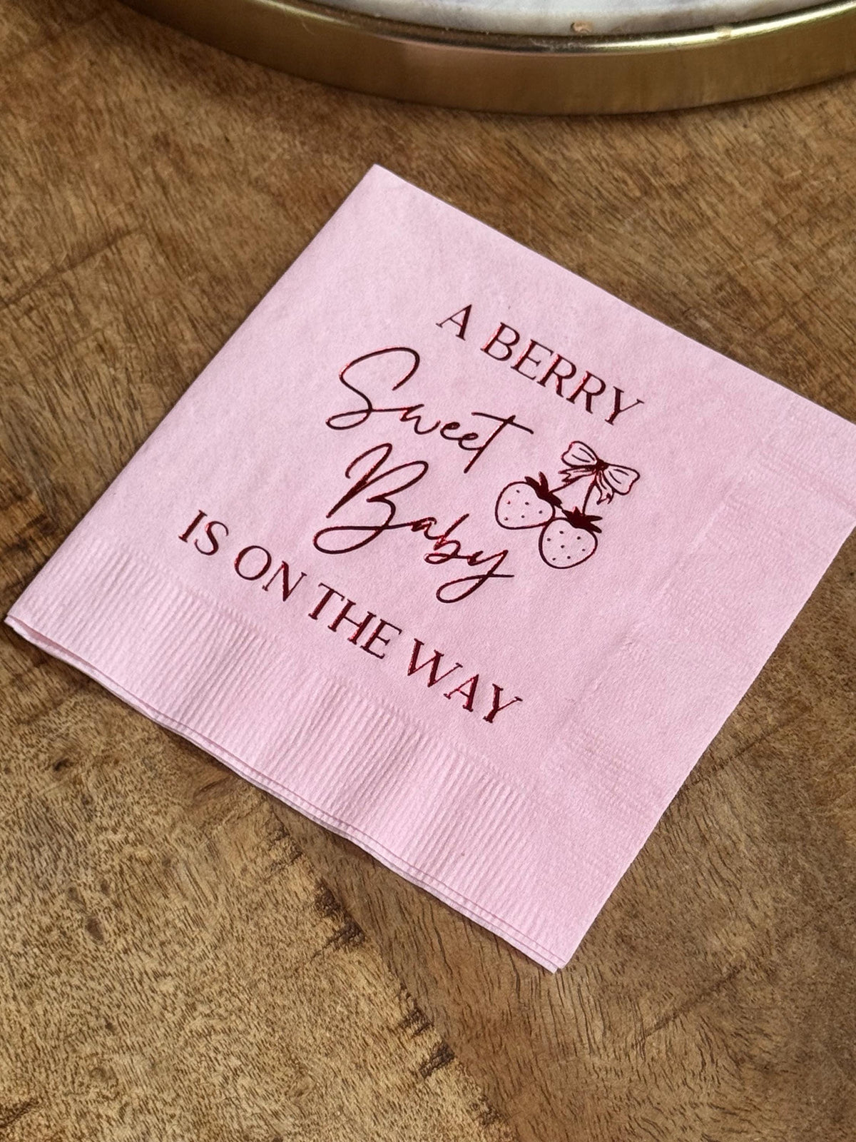 A single light pink cocktail napkin on a wooden surface with dark red text reading A Berry Sweet Baby Is On The Way and a strawberry illustration.