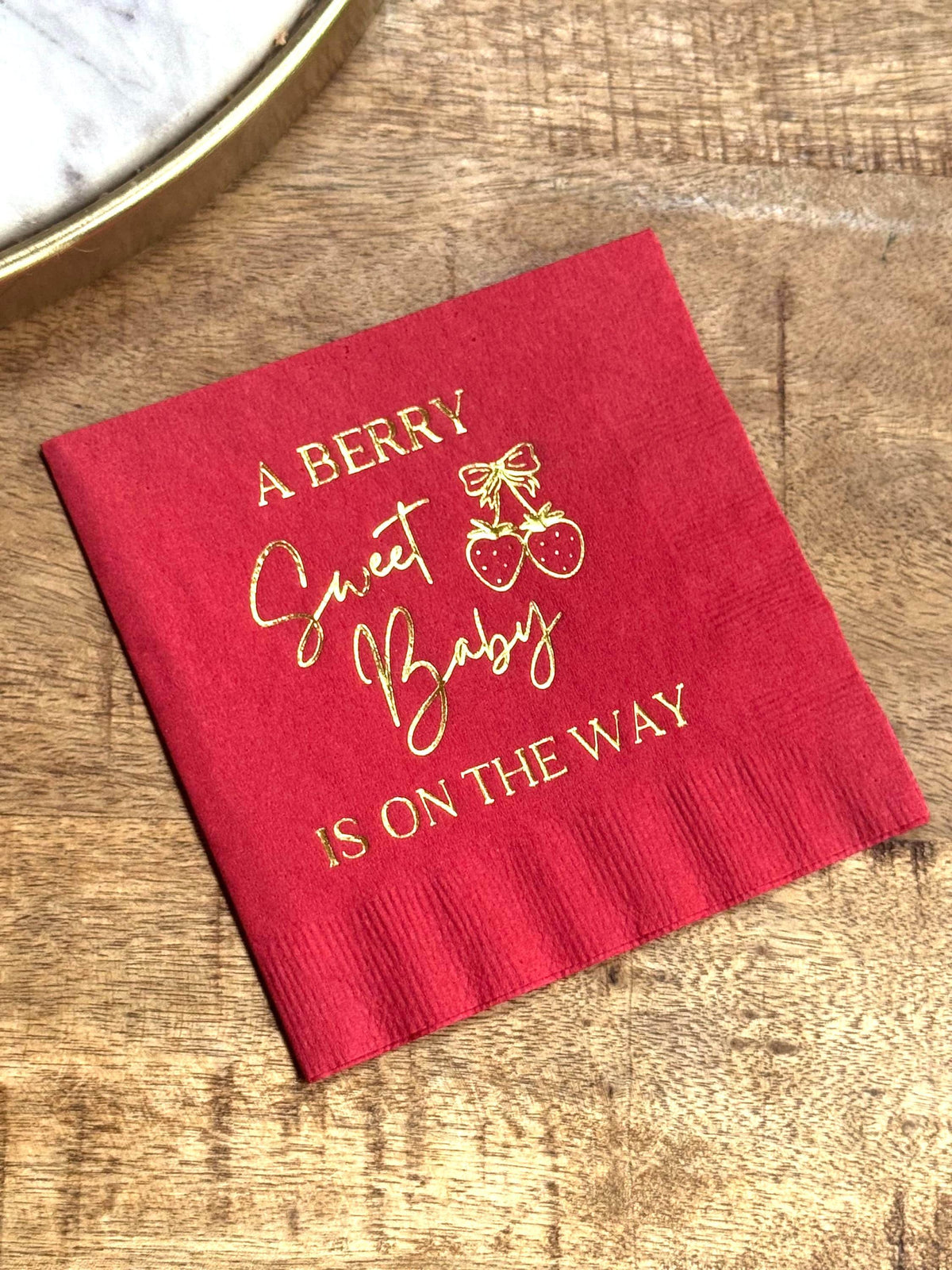 A red cocktail napkin on a wooden background with gold foil lettering reading A Berry Sweet Baby Is On The Way and a strawberry graphic.