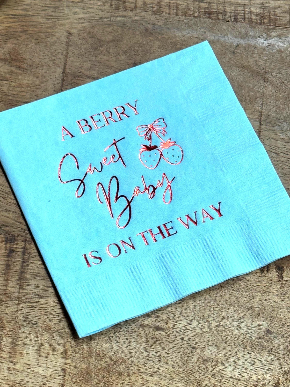 A light blue cocktail napkin on a wooden surface featuring rose gold foil stamped text reading A Berry Sweet Baby Is On The Way.