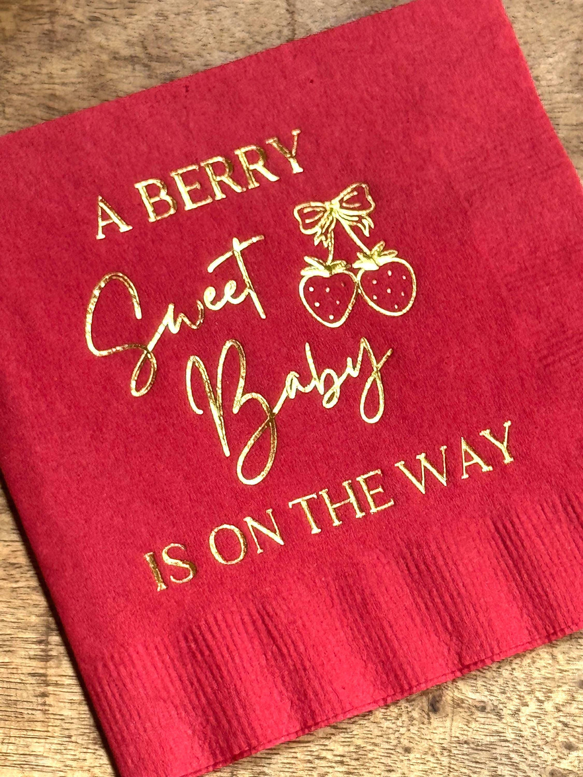 A close up view of a red cocktail napkin with gold foil stamping that reads A Berry Sweet Baby Is On The Way and two strawberries tied with a bow.