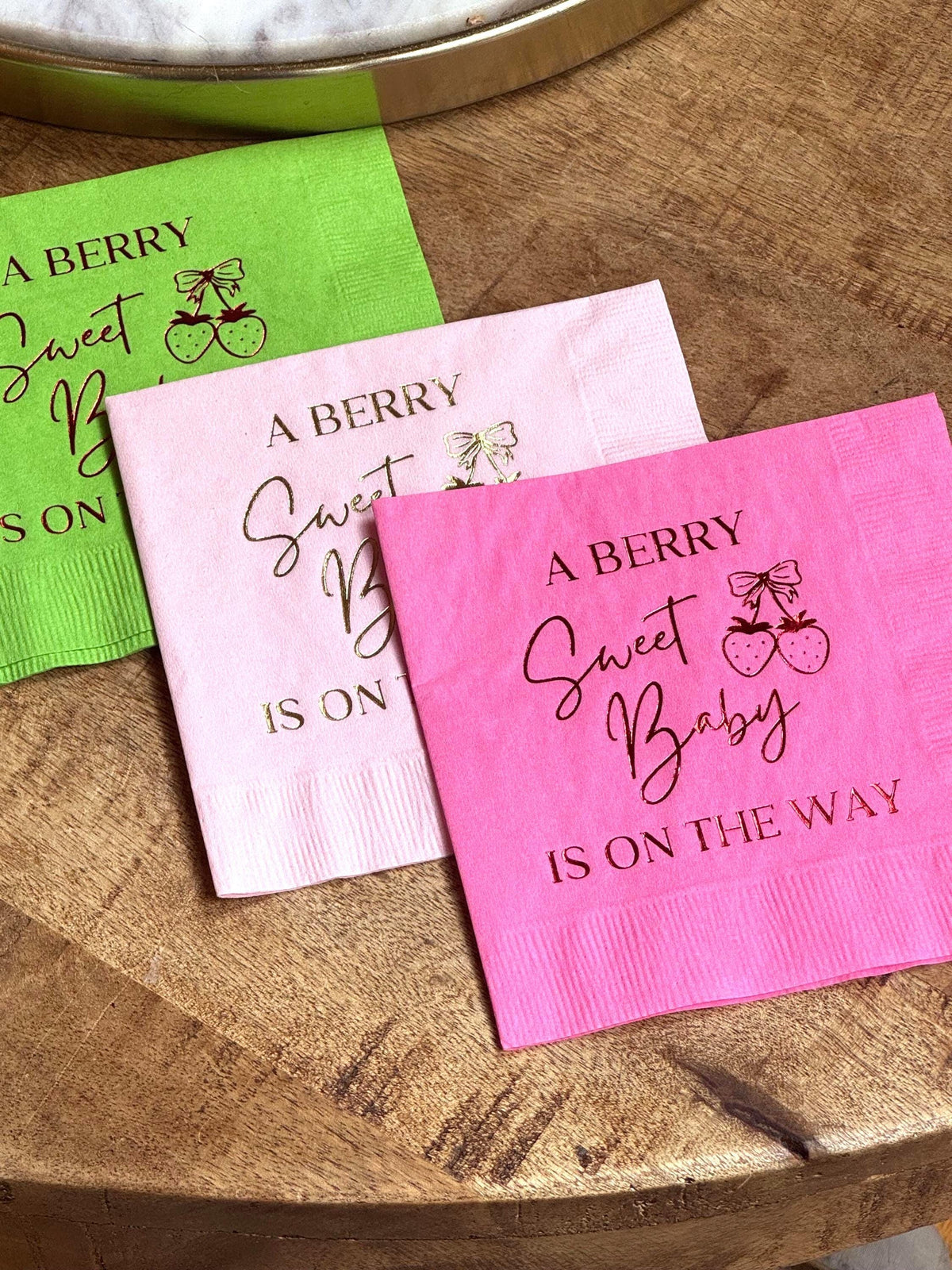 Three stacked cocktail napkins in green, light pink, and hot pink on a wooden table, all printed with the phrase A Berry Sweet Baby Is On The Way.