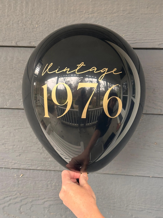 Black balloon with 'Vintage 1976' text held against a wooden surface