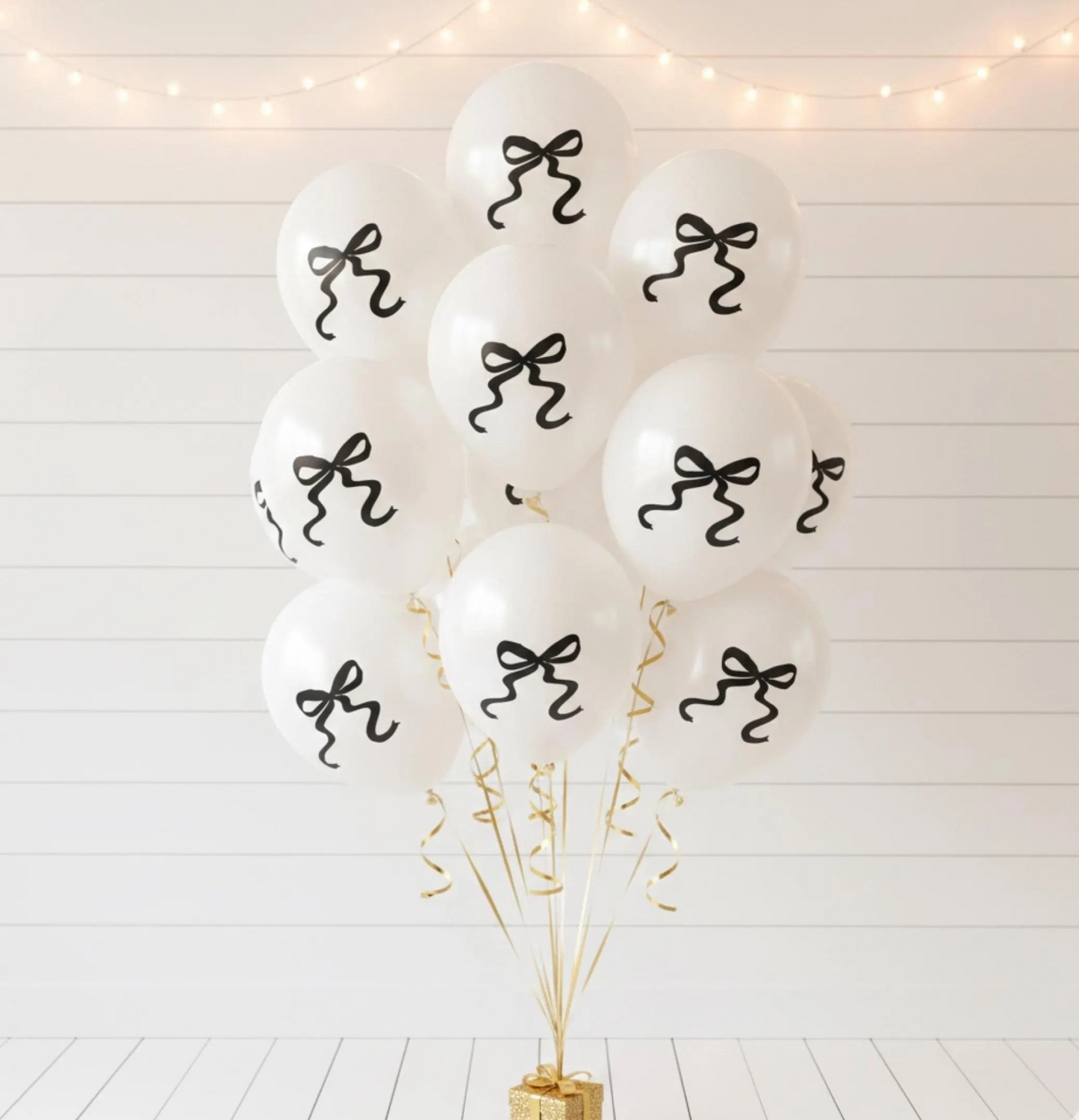 A cluster of white balloons with black bow designs, tied together with gold streamers and a gold base, set against a white paneled wall with warm string lights.