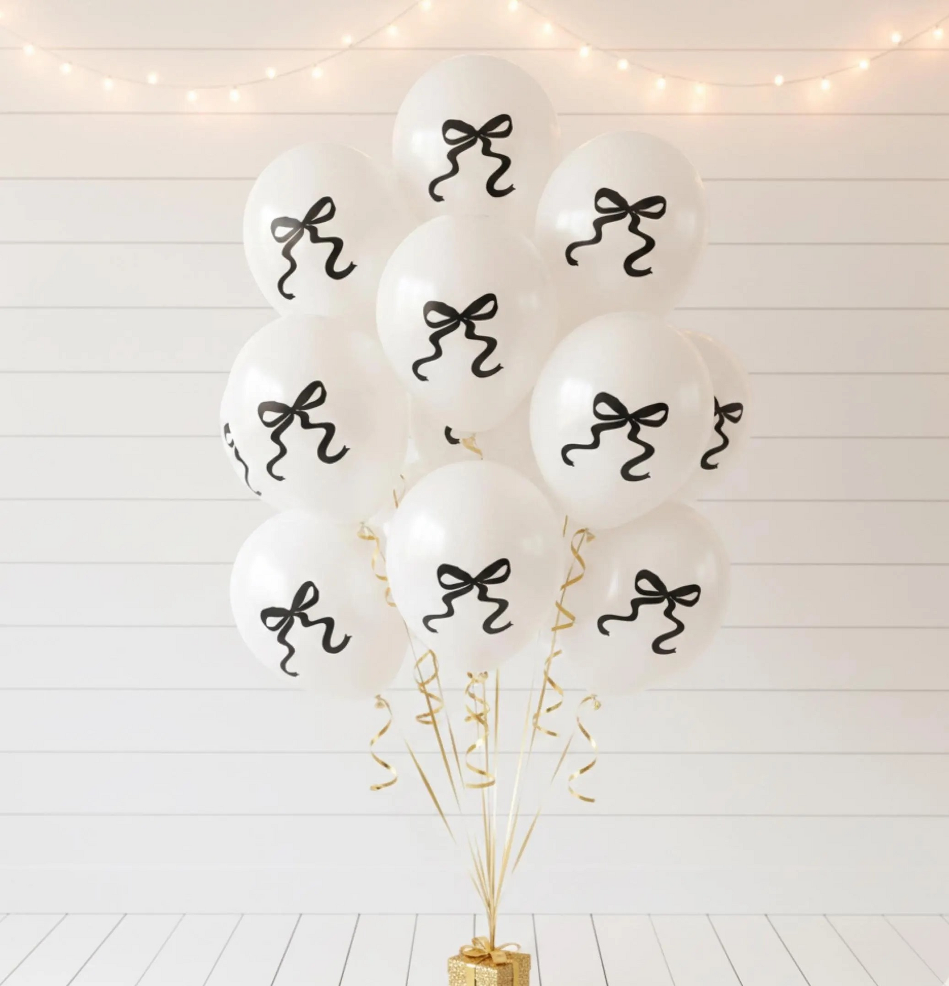 A cluster of white balloons with black bow designs, tied together with gold streamers and a gold base, set against a white paneled wall with warm string lights.