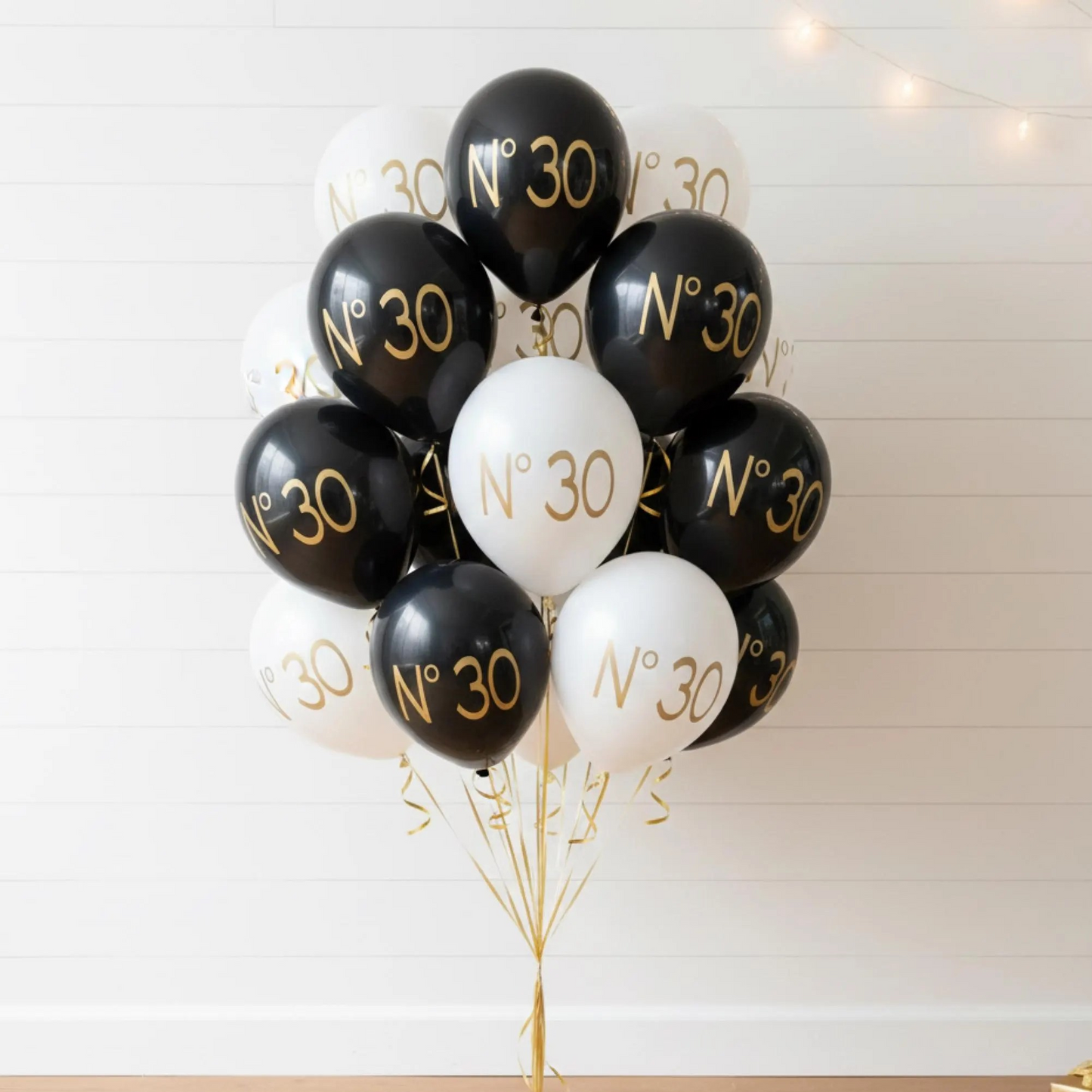 30th birthday balloons.  A balloon bouquet, featuring alternating black and white balloons printed with 'N° 30' in gold lettering, set against a white paneled wall with warm string lights. 30th Birthday decorations