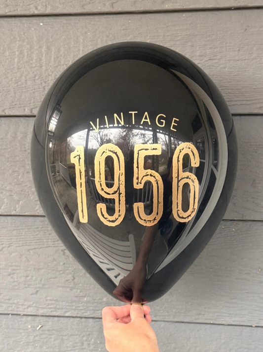 Black balloon with 'Vintage 1956' text held against a wooden surface