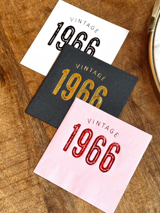 Three cocktail napkins with 'Vintage 1966' text in different colors on a wooden surface.