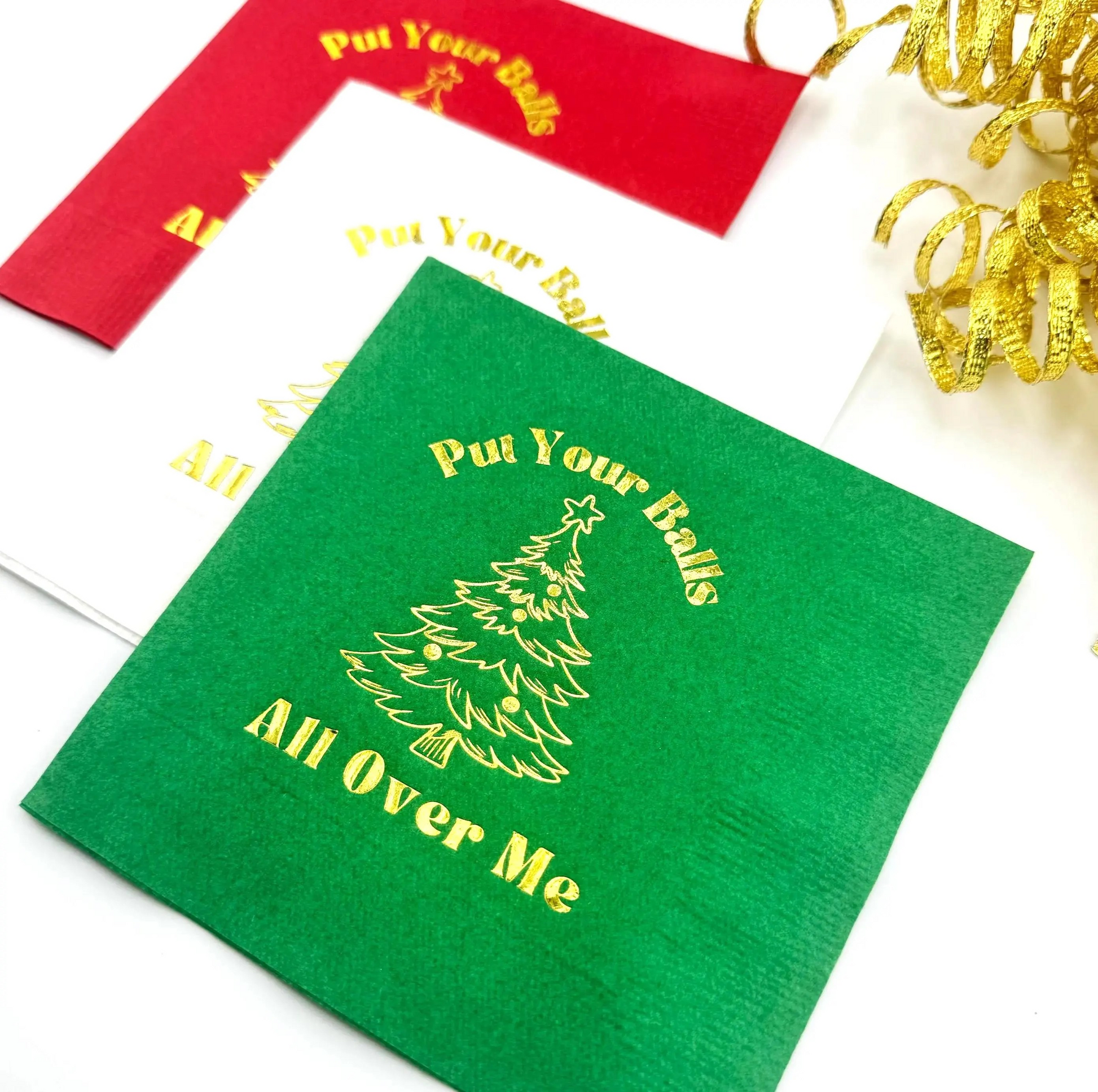 Green cocktail napkin with humorous text "Put your balls all over me" and a Christmas tree design, surrounded by red and white napkins.