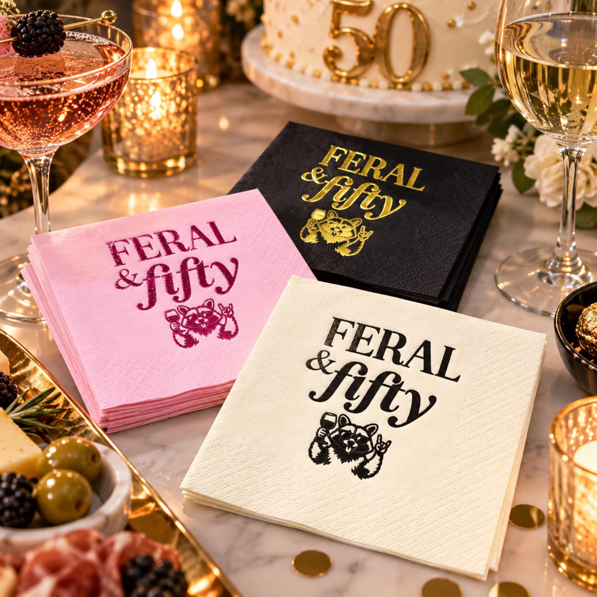 A party setting with food, drinks, and three stacks of cocktail napkins in black with gold foil, pink with magenta foil, and cream with black print, all reading Feral & fifty with a raccoon graphic.