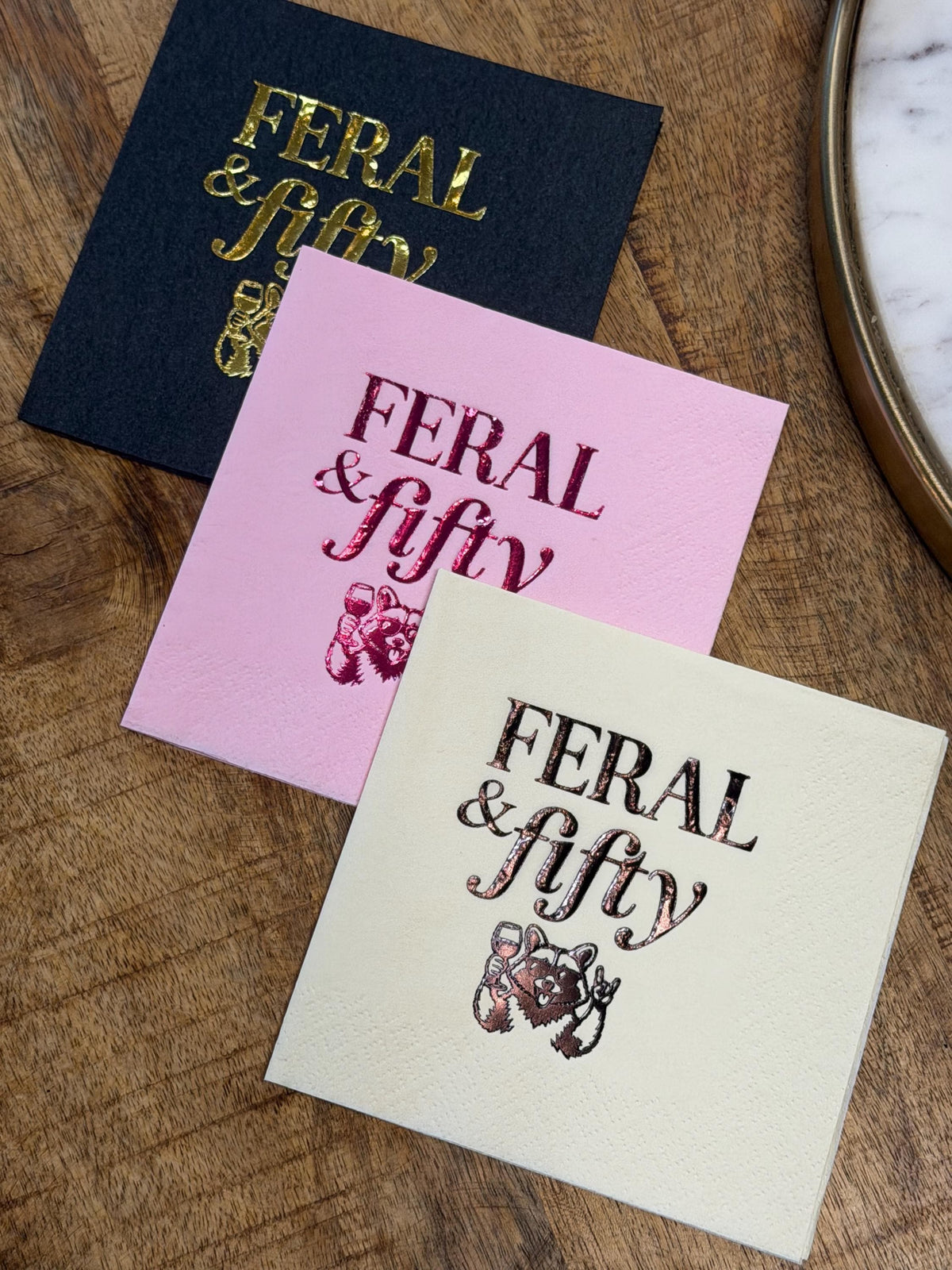 Three stacked square paper cocktail napkins in black with gold foil, pink with magenta foil, and cream with rose gold foil, printed with text reading FERAL and fifty and a raccoon graphic.
