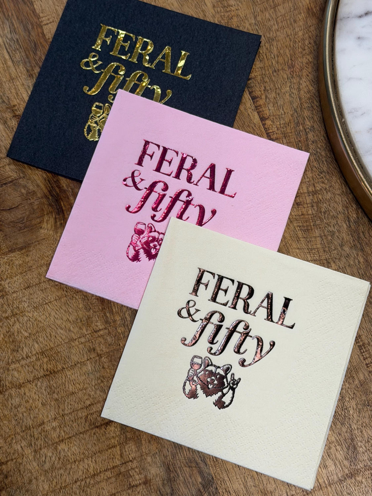 Close-up of three square paper beverage napkins featuring the text FERAL and fifty and an illustrated raccoon holding a wine glass, arranged on a wooden surface.