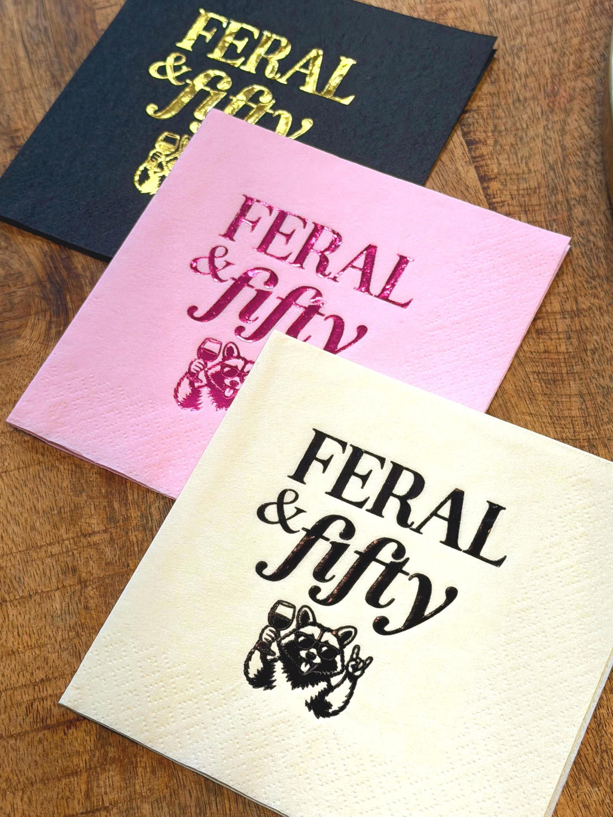 Three individual cocktail napkins laid on a wooden surface in black with gold foil, pink with magenta foil, and cream with black print, displaying a raccoon holding a wine glass and the text Feral & fifty.