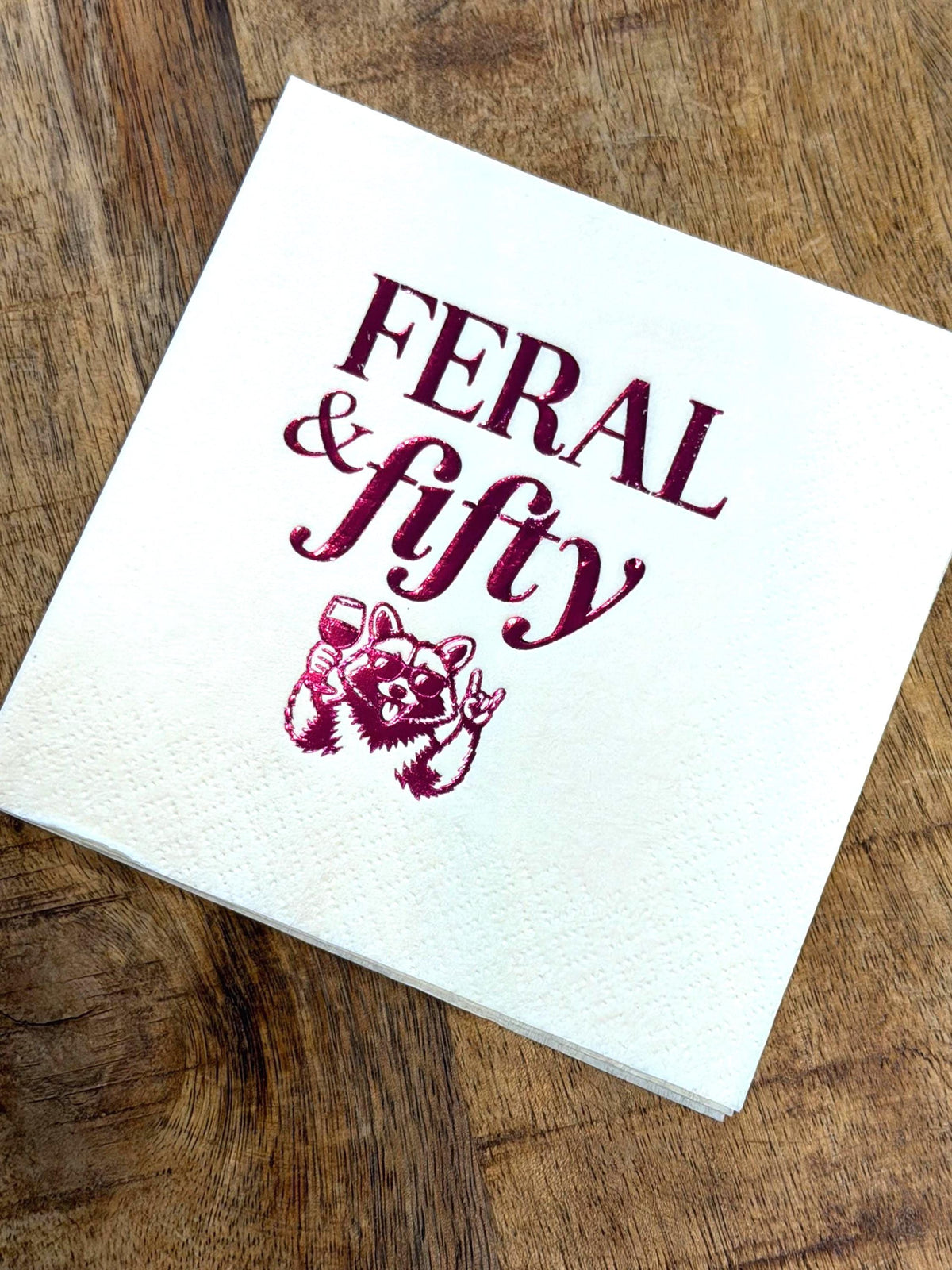 A single cream cocktail napkin on a wooden background featuring a raccoon in sunglasses holding a wine glass with the text Feral & fifty printed in dark red.