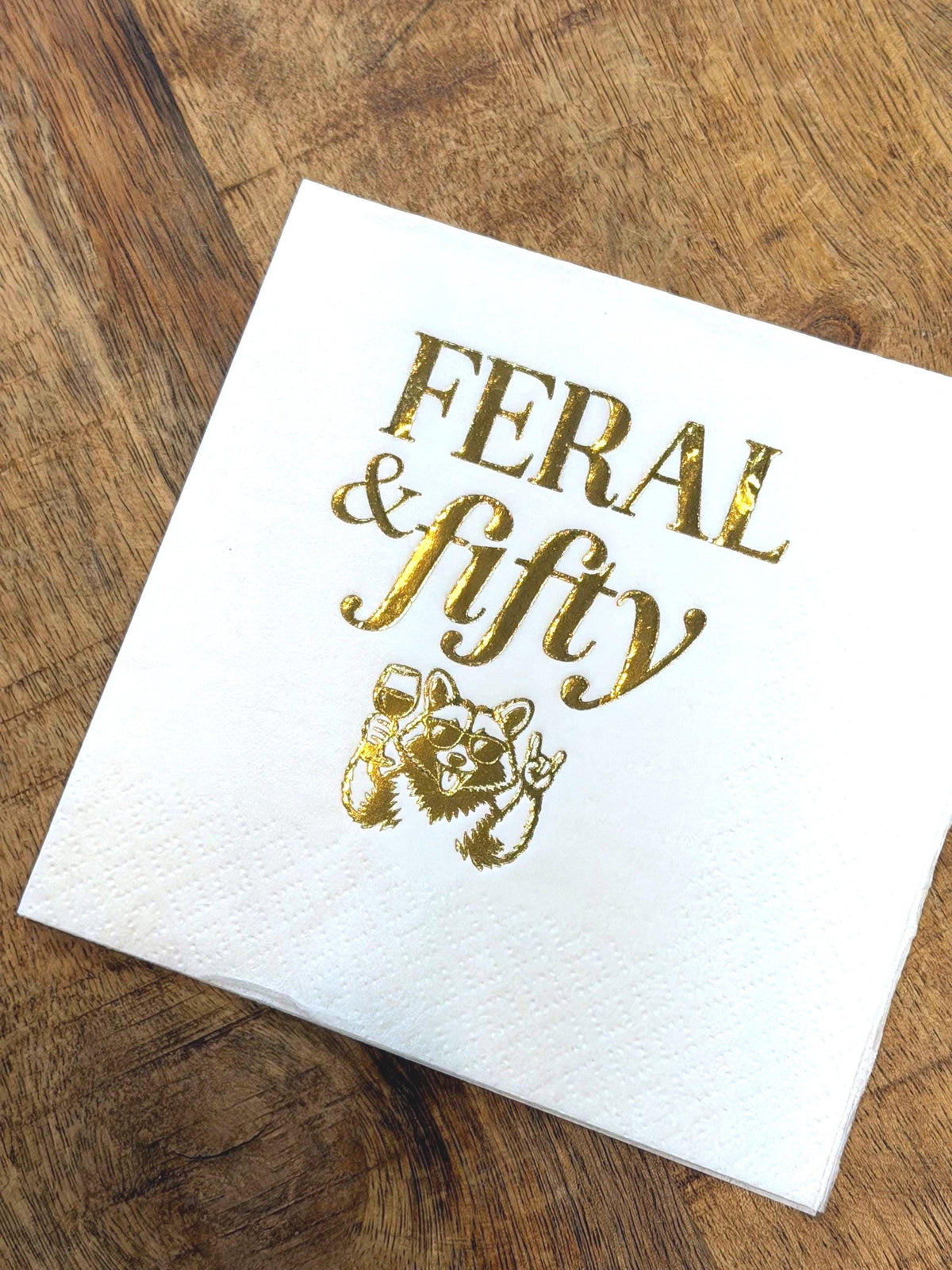 A single white cocktail napkin on a wooden surface with shiny gold foil text reading Feral & fifty above an illustration of a raccoon holding a wine glass.
