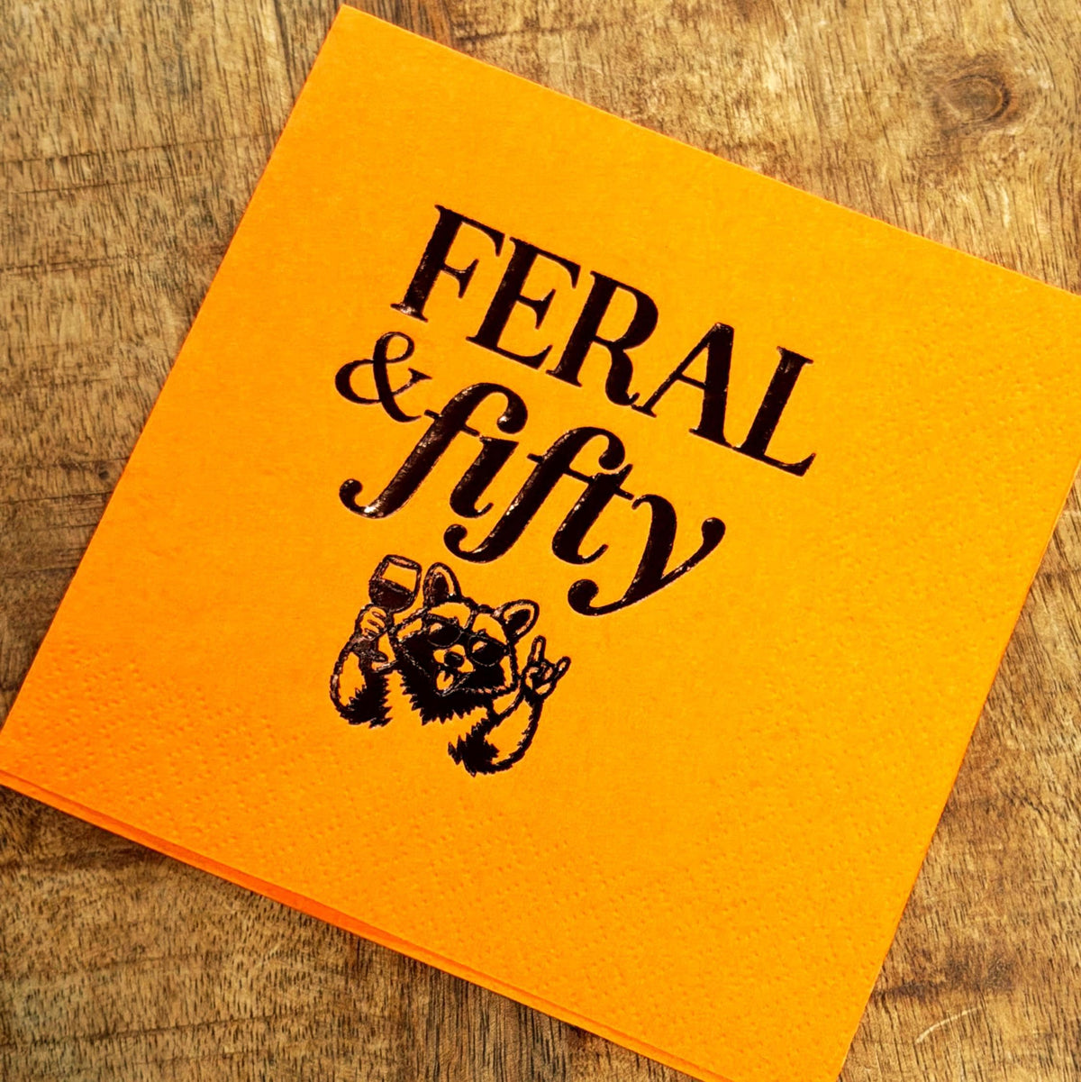 A single bright orange cocktail napkin on a wooden surface featuring black print of the text Feral & fifty and a raccoon holding a wine glass.
