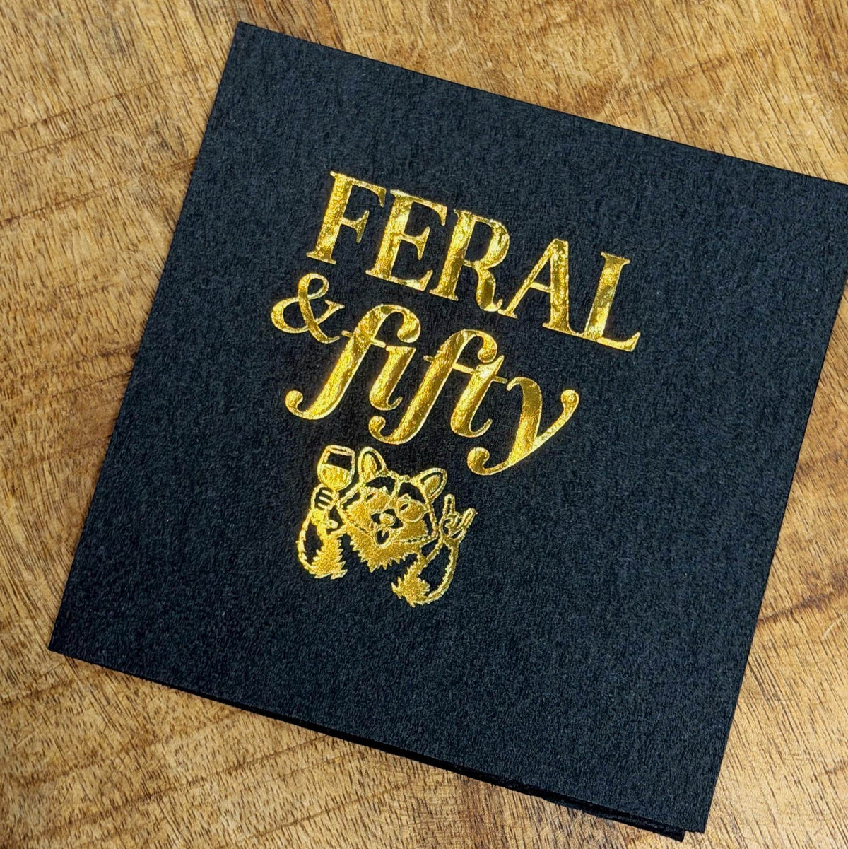 A single black cocktail napkin on a wooden surface with shiny gold foil lettering that reads Feral & fifty above a raccoon graphic.