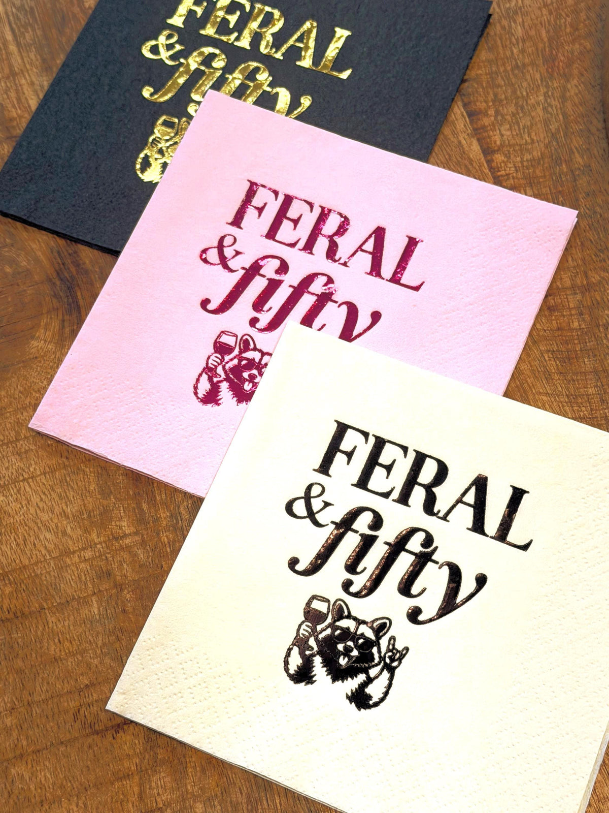 Three cocktail napkins overlapping on a wooden table, featuring black with gold foil, pink with magenta foil, and cream with black print, all with the Feral & fifty raccoon design.