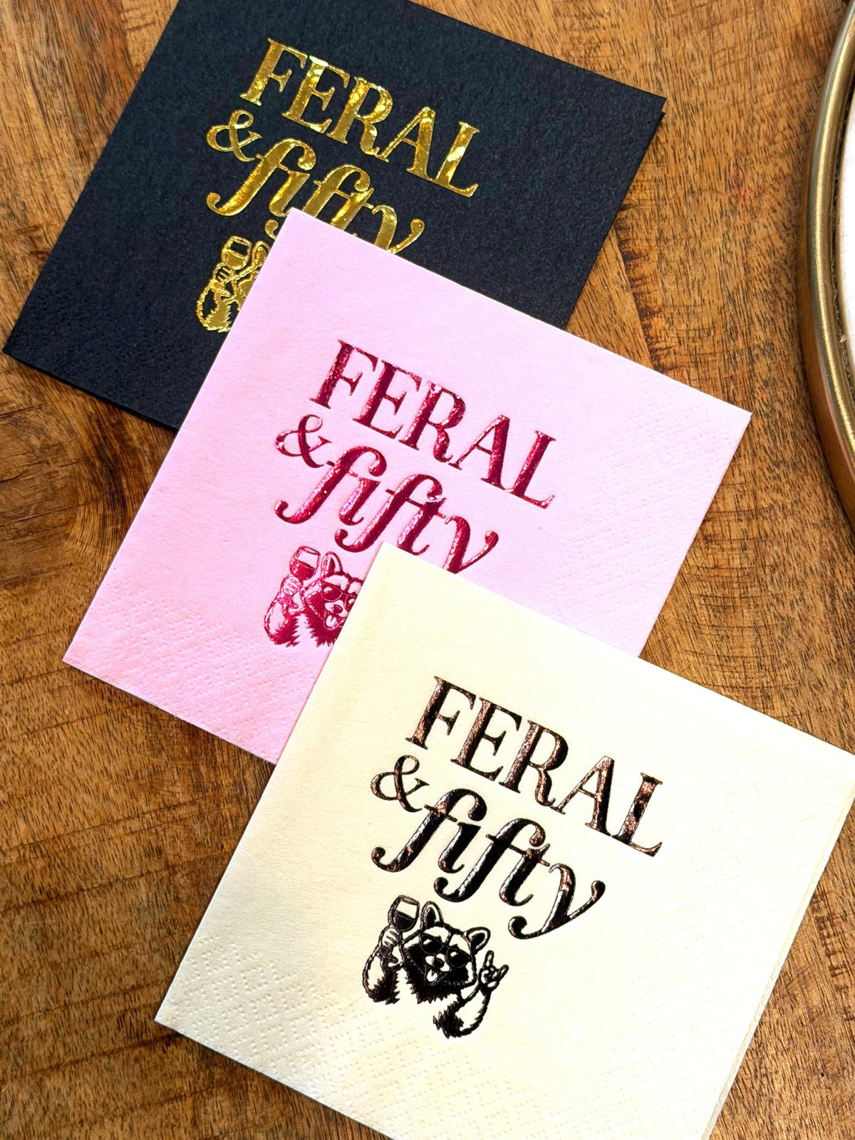 Three cocktail napkins displayed diagonally on a wooden surface in black with gold foil, pink with magenta foil, and cream with black print, showing the Feral & fifty raccoon design.