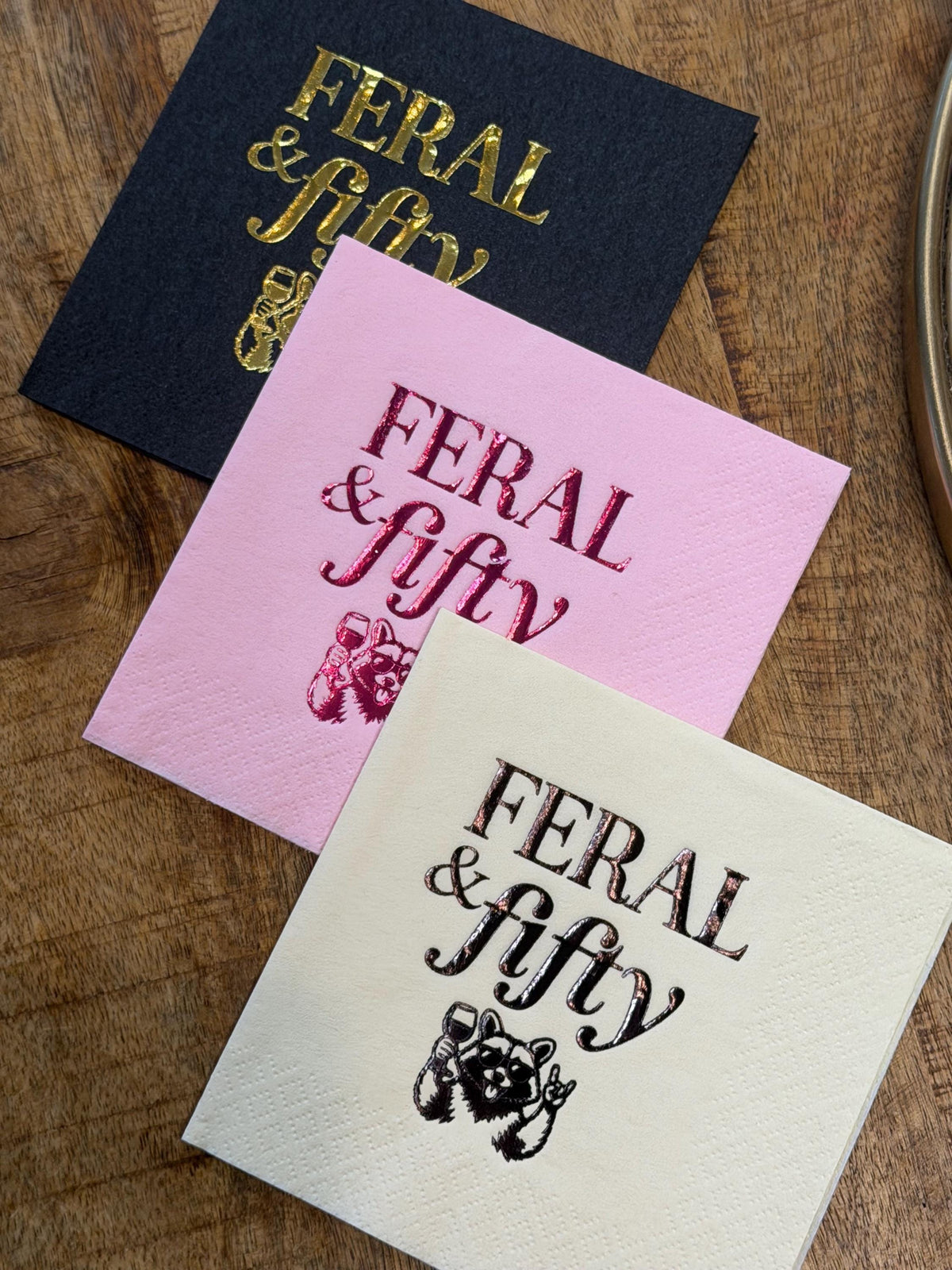 Three cocktail napkins angled on a wooden background in black with gold foil, pink with magenta foil, and cream with black print, featuring the Feral & fifty raccoon illustration.