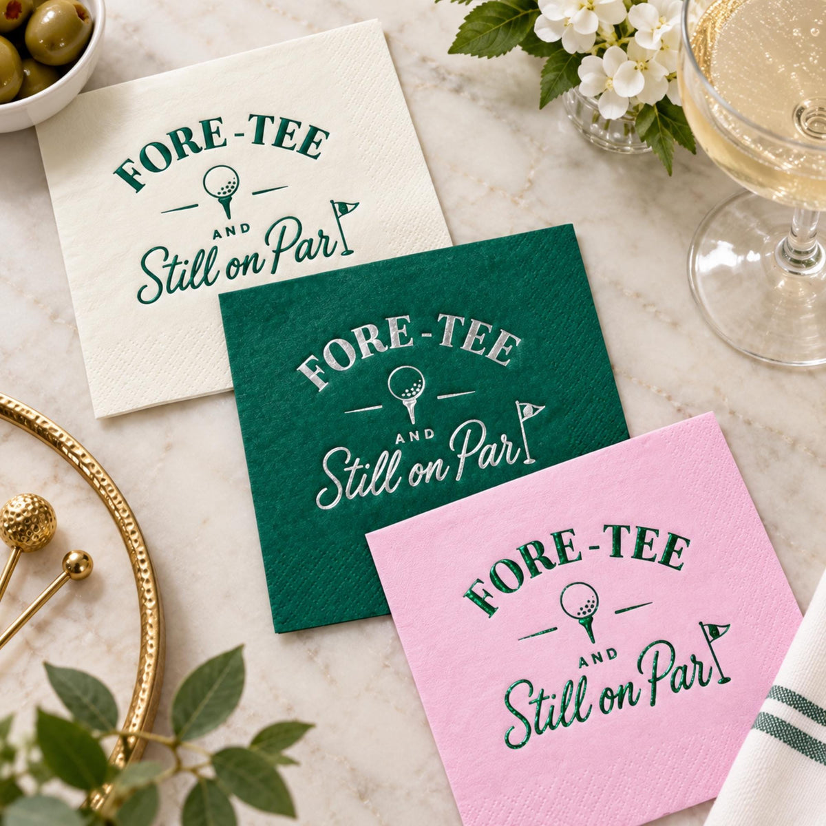 White, green, and pink cocktail napkins lie stacked on a marble surface next to a glass of champagne, olives, and a golf tee tray.
