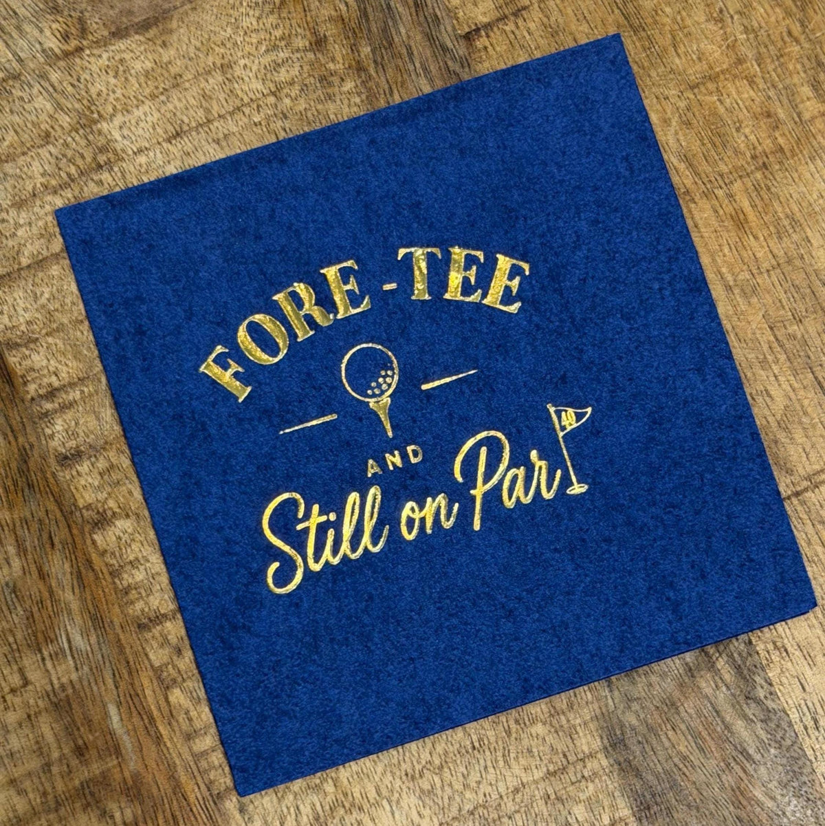 A dark blue paper cocktail napkin rests on a wooden surface, featuring gold foil stamping that reads FORE-TEE AND Still on Par with golf graphics.