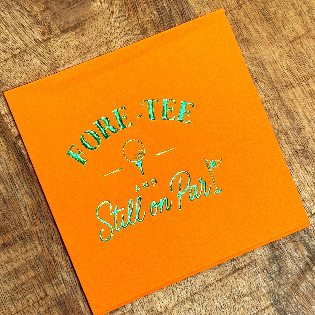 An orange paper cocktail napkin rests on a wooden tabletop, featuring metallic green foil stamping with the text FORE-TEE AND Still on Par and golf icons.